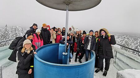 eine Gruppe junger Menschen steht auf einem Aussichtsturm in einer Winterlandschaft und lächelt von dort aus in die Kamera