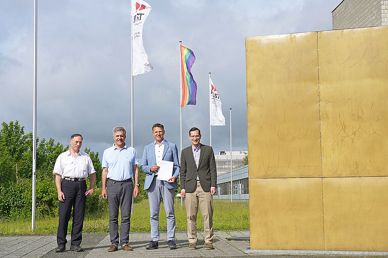 Vier Männer stehen neben einem großen goldenen Würfel, einem Kunstwerk am Bau, vor wehenden Flaggen, darunter die Regenbogenflagge und halten ein Blatt Papier hoch - lächelnd.