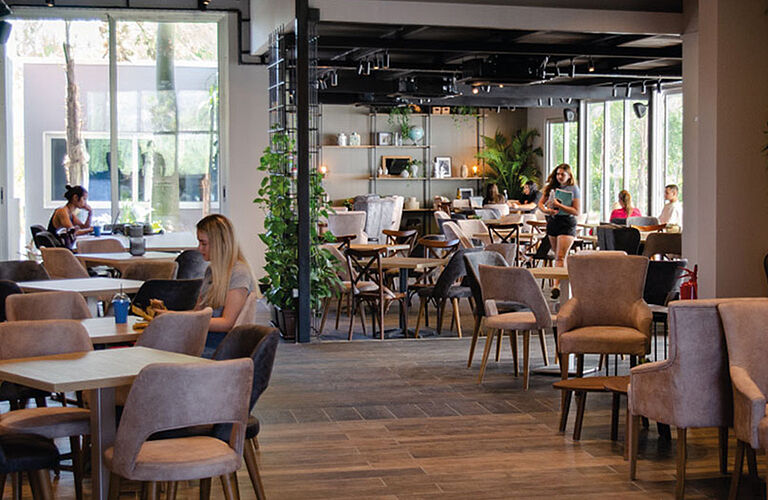 Blick in eine Cafeteria mit Naturoptikmöbeln, einzelnen Grünpflanzen und Studierenden