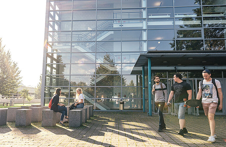 Studierende sitzen auf Bänke und gehen in Gebäude mit Glasfassade