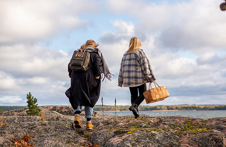 Zwei junge Frauen von hinten zu sehen, wie sie, augenscheinlich im Herbst, über eine steinige Küste ans Wasser gehen.