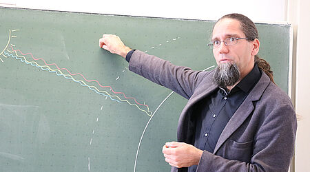 Prof. Johannes Gulden im dunklen Anzug steht vor einer grünen Tafel, auf der Sonne und Erde mit Kreide abgebildet sind