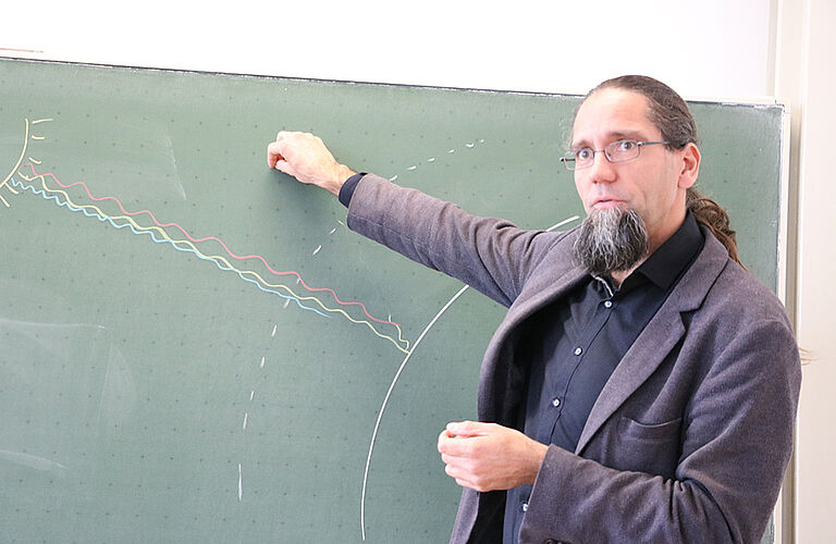 Prof. Johannes Gulden im dunklen Anzug steht vor einer grünen Tafel, auf der Sonne und Erde mit Kreide abgebildet sind