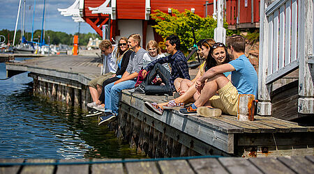 9 junge Personen in kurzen Hosen und T-Shirst sitzen an einem Steg mit Segelbooten im Hintergrund