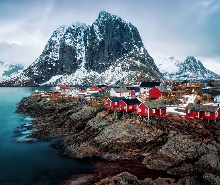 Klassische rote Norwegenhäuschen vor eisbedeckten Bergen an einer Klippe ins Meer