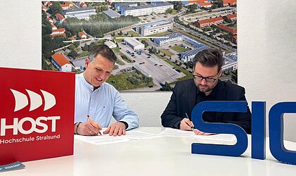 Zwei Männer unterschreiben jeweils ein Papier an einem Schreibtisch sitzend