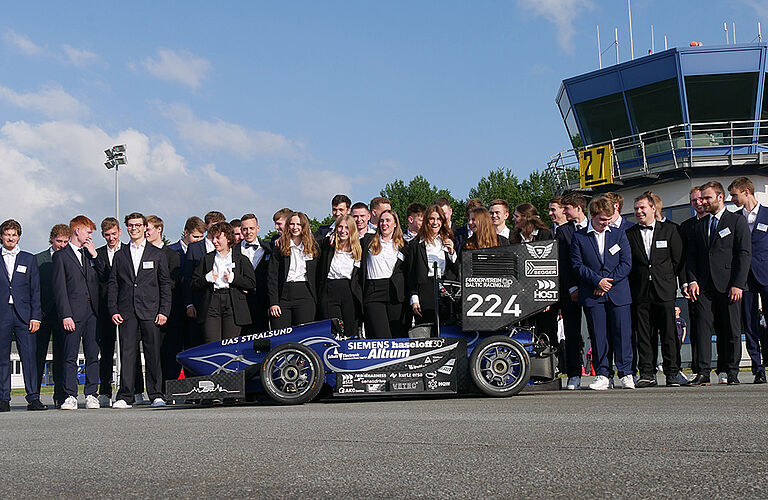 Gruppenfoto des Batic Racing Teams, in blauen und schwarzen Anzügen, mit ihrem ersten voll-elektrischen Formel-Student-Rennwagen auf dem Flughafen in Barth.