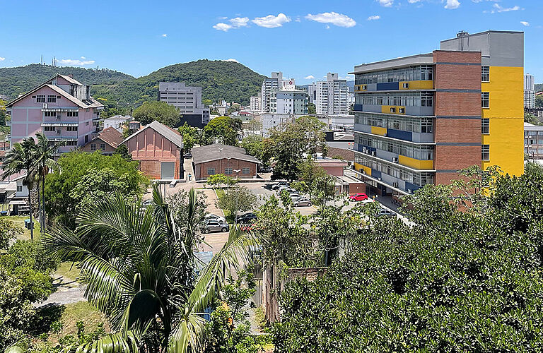 Luftaufnahme der Campusgebäude der Universidade Regional de Blumenau mit Palmen im Vordergrund und begrünten Bergen im Hintergrund