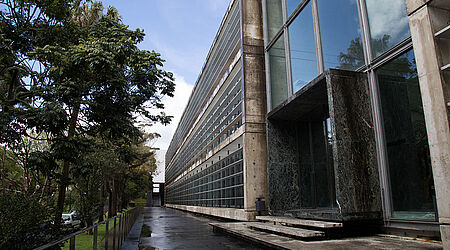 Blick auf einen Gehweg. Links davon ist ein bewaldeter Grünstreifen. Rechts davon befindet sich ein futuristisch aussehendes graues Haus mit anteiligen Glasfassade.