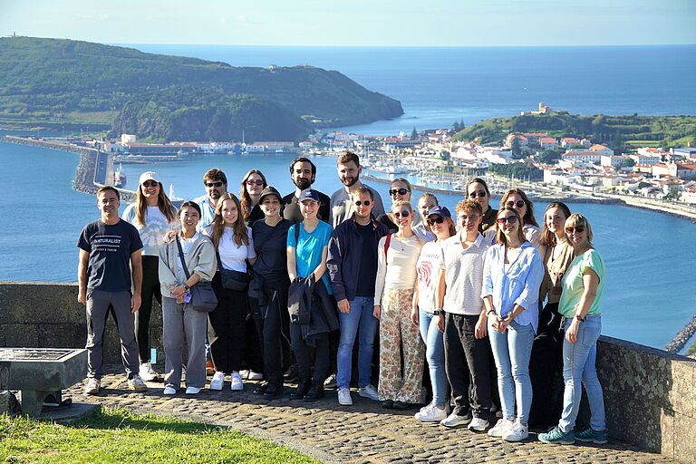 ein Gruppenbild mit rund 20 lächelnden Personen vor einer idyllischen Küstenlandschaft. 