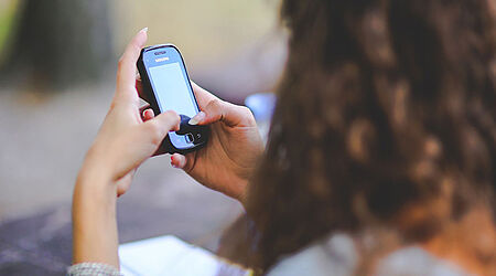 verschwommene Aufnahme über die Schulter einer Frau von hinten, die ein Smartphone in den Händen hält