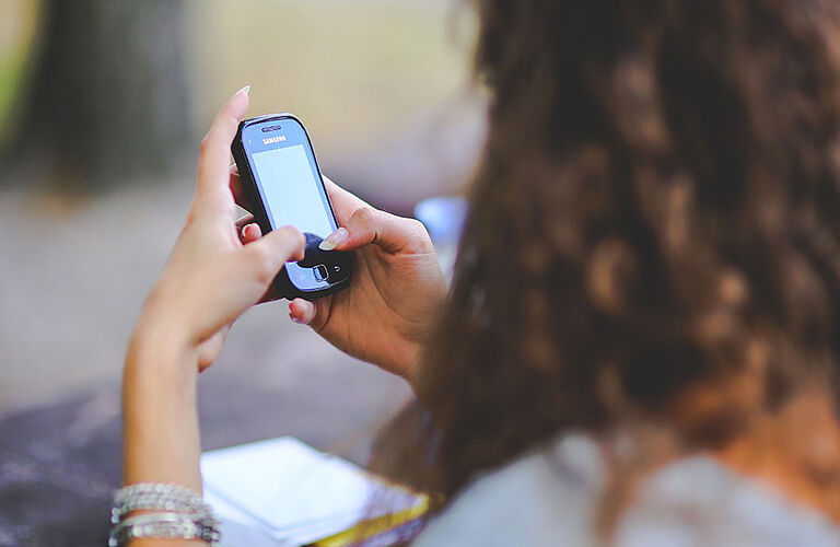 verschwommene Aufnahme über die Schulter einer Frau von hinten, die ein Smartphone in den Händen hält