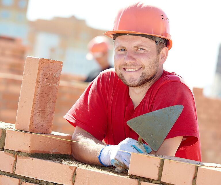 ein lächelnder Maurer mit Helm und rotem T-Shirt, sich mit Maurerkelle auf eine Ziegelmauer stützend