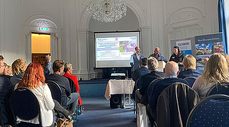 Mehrere Menschen sitzen in einem barock verzierten Saal bei einer Präsentation von Angehörigen der HOST.