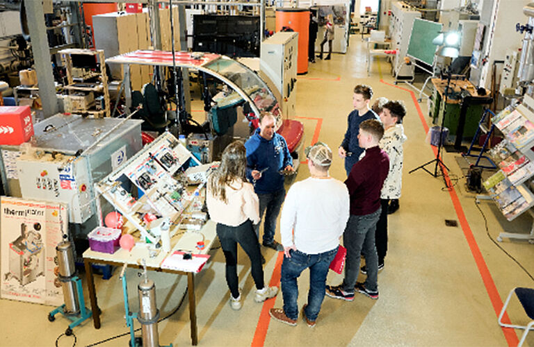 eine Gruppe junger Menschen mit Andre Sklarow der ihnen Dinge in einem großen Labor mit vielen technischen Aufbauten erklärt