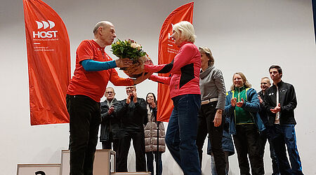 Prof. Wolfgang Schikorr werden Blumen von einer Frau überreicht. Sie stehen vor einem Siegertreppchen und sind umrahmt von applaudierenden Menschen.