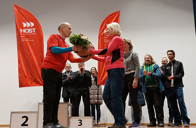 Prof. Wolfgang Schikorr werden Blumen von einer Frau überreicht. Sie stehen vor einem Siegertreppchen und sind umrahmt von applaudierenden Menschen.