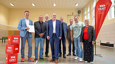 Eine Menschengruppe steht in der Sporthalle der HOST neben einer roten HOST-Flagge.