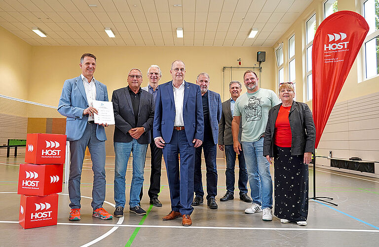 Eine Menschengruppe steht in der Sporthalle der HOST neben einer roten HOST-Flagge.