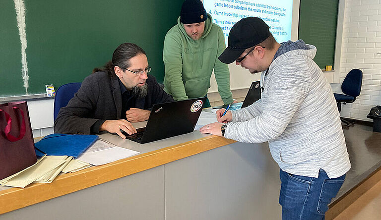 Ein Mann sitzt an einem Pult an einem Laptop, rechts neben ihm zwei jüngere Männer die mit auf Laptop oder Unterlagen schauen. 