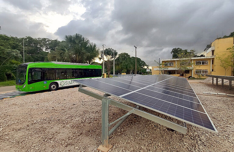 Nachaufnahme von Solarplatten mit Palmen und grünem Bus im Hintergrund