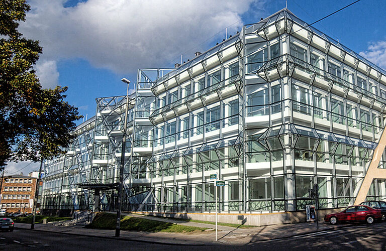 Ein großes Eckhaus, eines Häuserblocks, mit dominanter Glasfassade bei strahlend blauem Himmel.