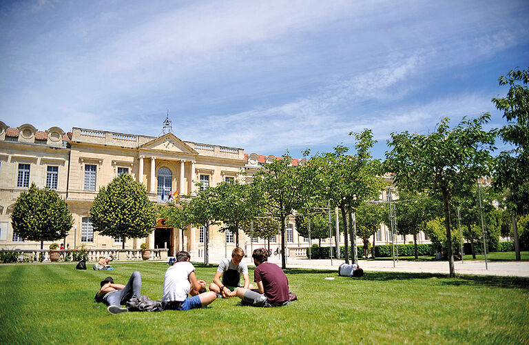 Großes Uni-Gebäude mit grüner Wiese und sitzenden, jungen Menschen.