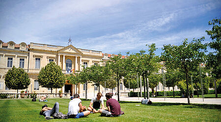 Großes Uni-Gebäude mit grüner Wiese und sitzenden, jungen Menschen.