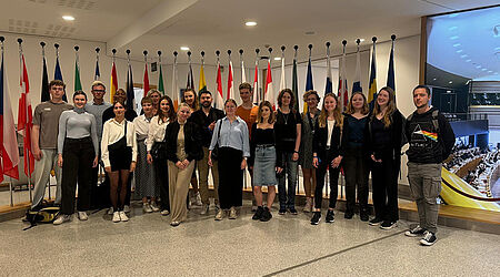 19 Studierende mit Prof. Claudia Danker beim Gruppenbild der Brüssel-Exkursion 2024 in einem Marmorsaal mit mehreren aufgestellten Flaggen unterschiedlicher EU-Länder