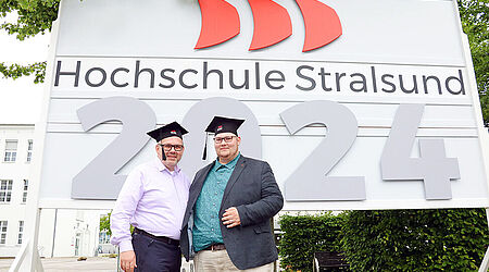 Zwei Männer mit Absolventenkappen stehen vor einem Hochschul-Schild mit einer großen 2024.