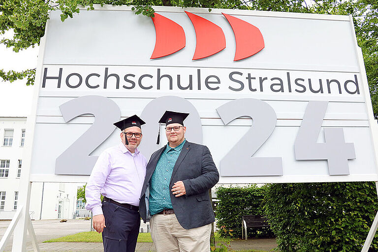 Zwei Männer mit Absolventenkappen stehen vor einem Hochschul-Schild mit einer großen 2024.