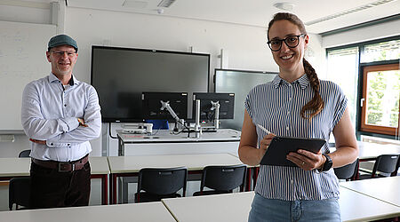 Prof. Gero Szepannek, im weißen Hemd, und Dr. Rabea Aschenbruck stehen in einem Seminarraum der HOST und lächeln in die Kamera.