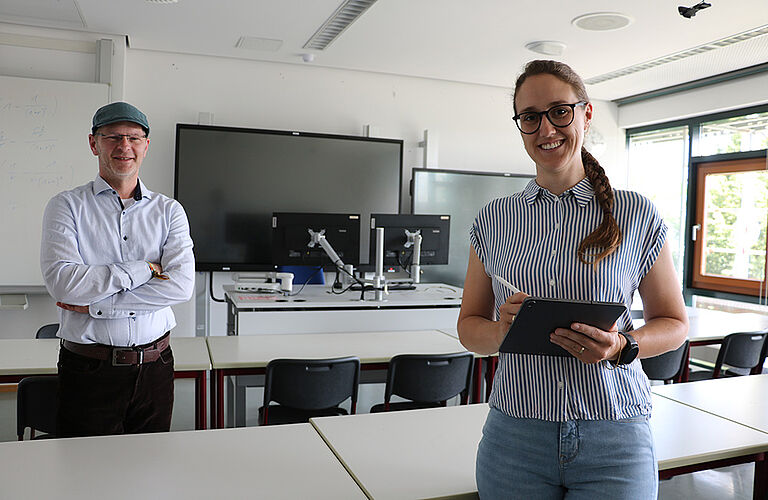 Prof. Gero Szepannek, im weißen Hemd, und Dr. Rabea Aschenbruck stehen in einem Seminarraum der HOST und lächeln in die Kamera.