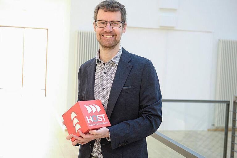 Prof. Jens Mohrenweiser, in blauem Sakko, hält einen roten HOST-Würfel in der Hand und steht lächelnd vor einem Treppengeländer