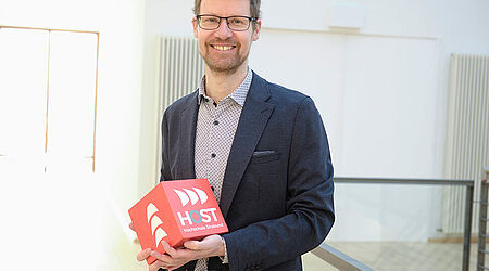 Prof. Jens Mohrenweiser, in a blue jacket, holds a red HOST cube in his hand and stands smiling in front of a stair railing