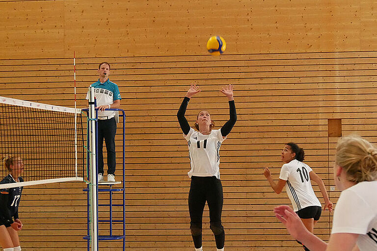 eine Volleyballerin springt vor dem Netz, um den Ball zu stellen und 2 weitere Spielerinnen machen sich zum Angriff bereit und bewegen sich auf das Netz zu