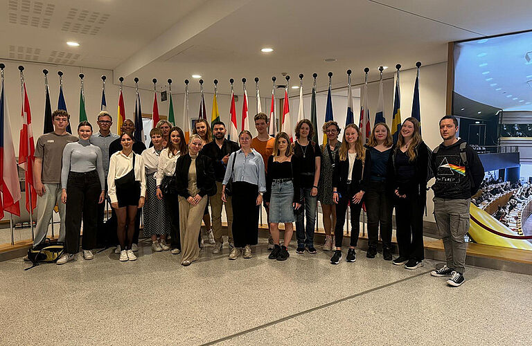 19 Studierende mit Prof. Claudia Danker beim Gruppenbild der Brüssel-Exkursion 2024 in einem Marmorsaal mit mehreren aufgestellten Flaggen unterschiedlicher EU-Länder