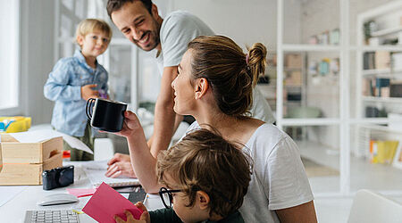 Eine Familie, mit zwei Kindern, sitzt an einem Tisch, in einem hellen, lichtdurchfluteten Raum. Die Eltern trinken Kaffe und lächeln sich gegenseitig an.