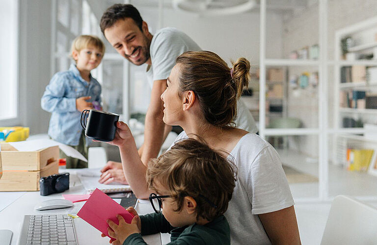 Eine Familie, mit zwei Kindern, sitzt an einem Tisch, in einem hellen, lichtdurchfluteten Raum. Die Eltern trinken Kaffe und lächeln sich gegenseitig an.