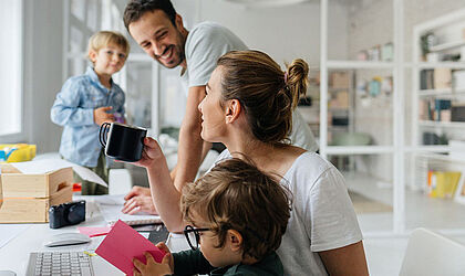 A family with two children is sitting at a table in a bright, light-flooded room. The parents are drinking coffee and smiling at each other.