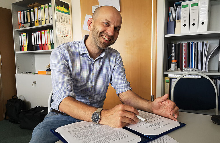 Prof. Normen Fuchs sitzt lächelnd an seinem Schreibtisch und sichtet Unterlagen