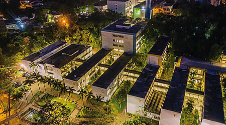 nächtliche Luftaufnahme mehrerer Gebäude der Universität Blumenau mit hellen Lichtern und Palmen