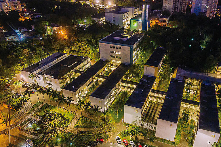 nächtliche Luftaufnahme mehrerer Gebäude der Universität Blumenau mit hellen Lichtern und Palmen