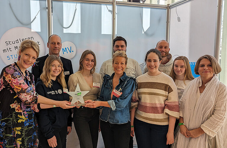 10 lächelnde Menschen schauen in die Kamera, einige halten einen sternförmigen Pokal (Stuzubi Star) zur Auszeichnung des besten Messestand auf Leipziger Messe.