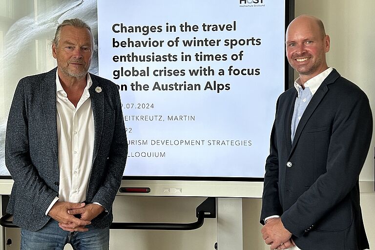 Zwei Männer stehen (leicht) lächelnd vor einem Bildschirm auf dem auf Englisch der Titel einer Abschlussarbeit zu lesen ist. "Changes in the travel behavior oft winter sports enthusiasts in times of gloabl crisis with a focus of the Austrian Alps"