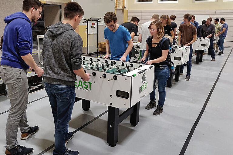 4 weiße, in Reihe gestellte Tischkicker mit jeweils 4 Spielern in der Turnhalle der HOST mit Publikum