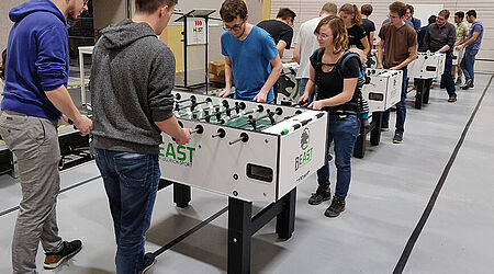 4 weiße, in Reihe gestellte Tischkicker mit jeweils 4 Spielern in der Turnhalle der HOST mit Publikum