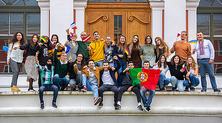 Eine Gruppe von internationalen Studierenden tummelt sich teils sitzende, teils stehend beim Fahnen wedeln vor dem Eingang eines weiß-roten Gebäudes