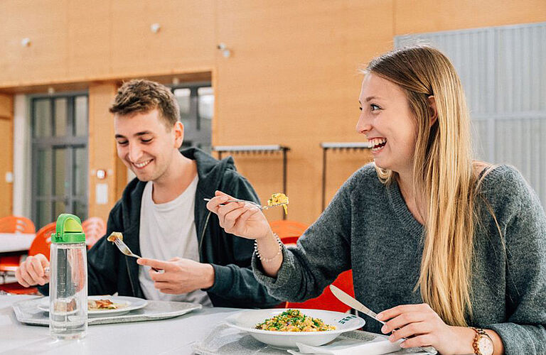 Nahaufnahme zweier lachenden Studierender, die in der HOST-Mensa sitzen und essen.