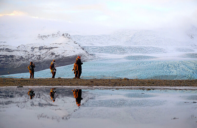 3 Personen auf Deich, die sich im Wasser spiegeln, vor eisiger Bergregion